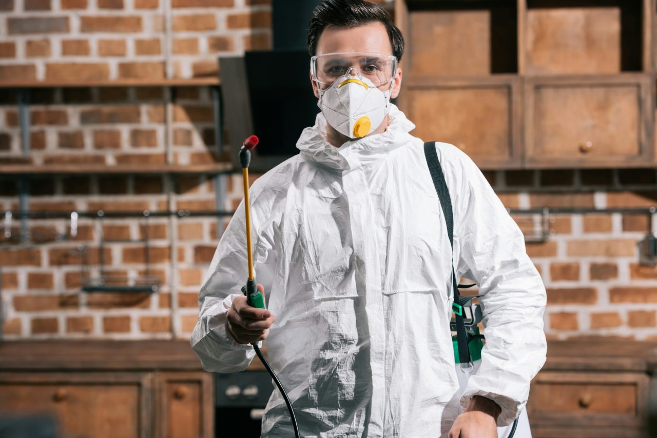 pest control worker standing with sprayer in kitchen and looking at camera